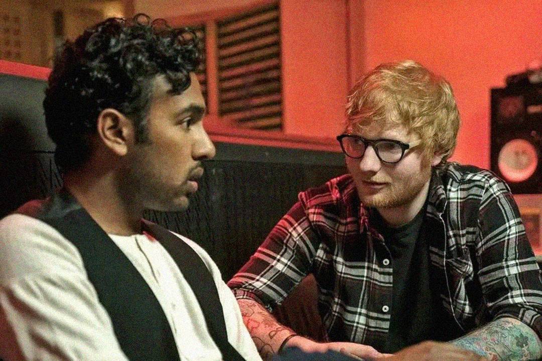 Two men sit closely together in a dimly lit room. One has curly hair and wears a white shirt with a black vest; the other has light hair, glasses, and wears a plaid shirt, looking at the first man while speaking.