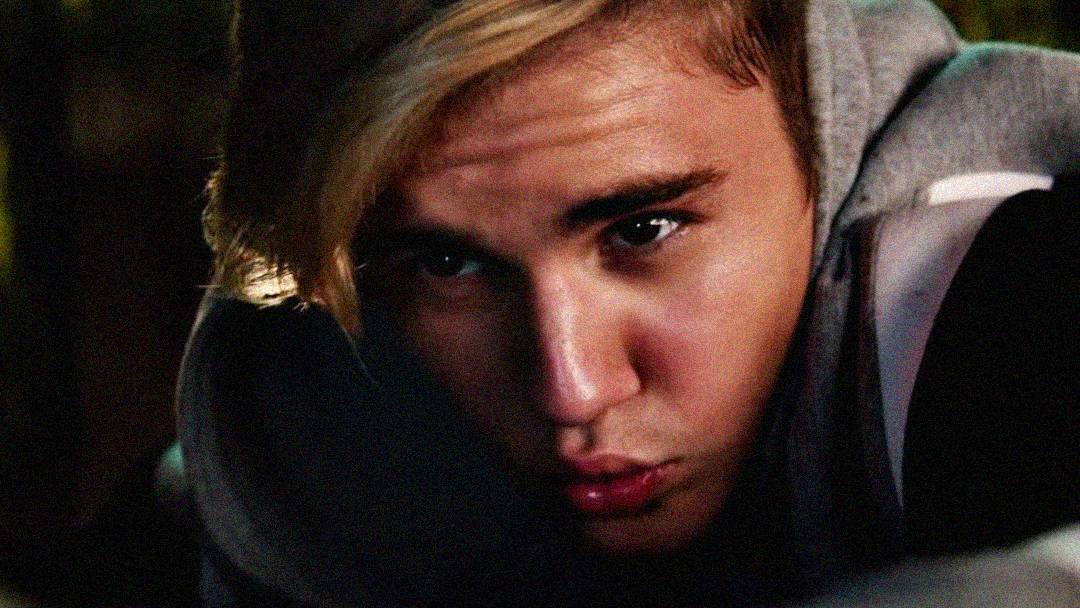 A young person with blonde hair and a gray hoodie is leaning forward, gazing directly at the camera with a serious expression and pursed lips. The background is dark and out of focus.