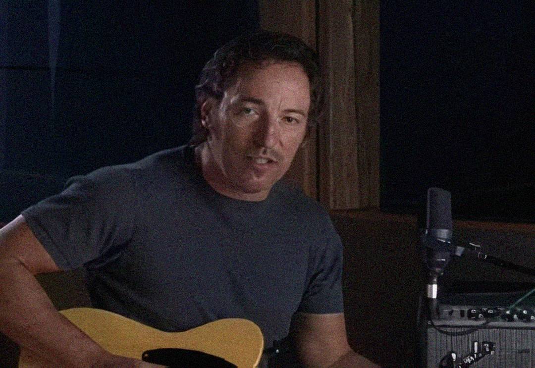 A man with wavy dark hair sits indoors, playing a yellow electric guitar. He wears a dark short-sleeve shirt and looks toward the camera. A microphone and amplifier are visible beside him.
