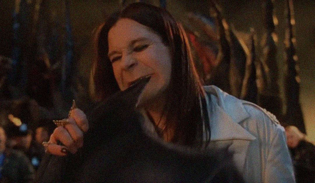 A person with long dark hair and a white jacket appears to bite the head of a black object in a dimly lit setting, with an intense facial expression.
