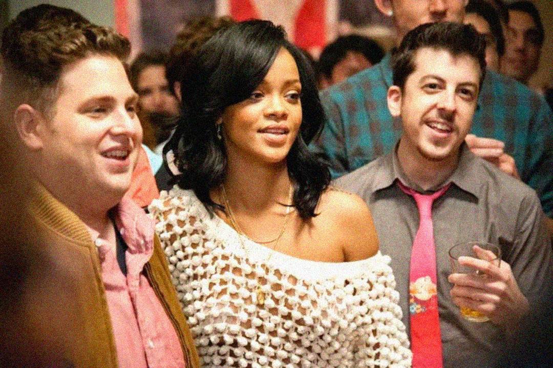A woman in a white, off-shoulder mesh top stands between two men at a crowded indoor social gathering. All three are smiling, and one man holds a drink. The atmosphere is lively and festive.