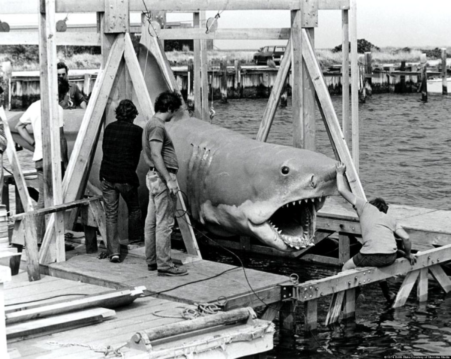A group of people stand on a dock beside a large mechanical shark, suspended by a wooden frame over the water. The scene appears to be behind the scenes of a film production.