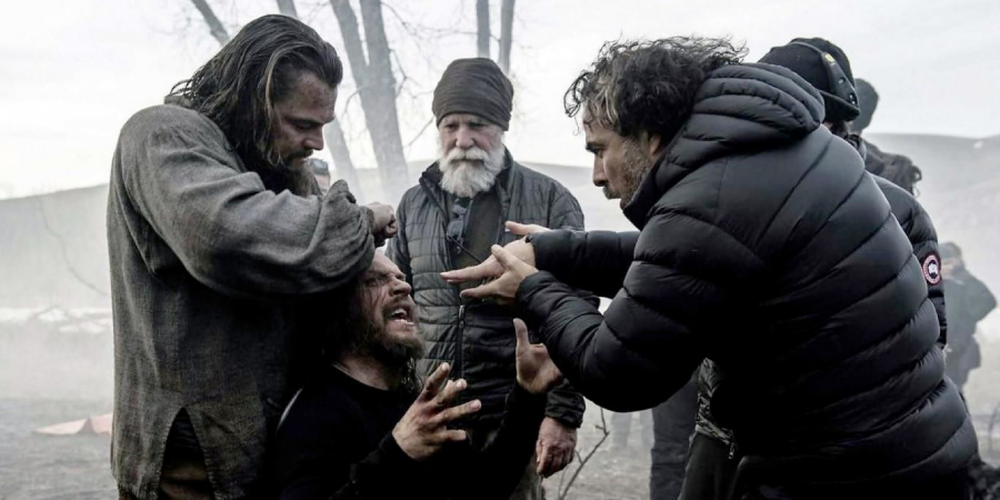 Three men are outside in a cold, foggy setting; one is kneeling and distressed, one is gripping his head, an older man stands behind them, and another man in a puffy jacket directs the scene passionately.