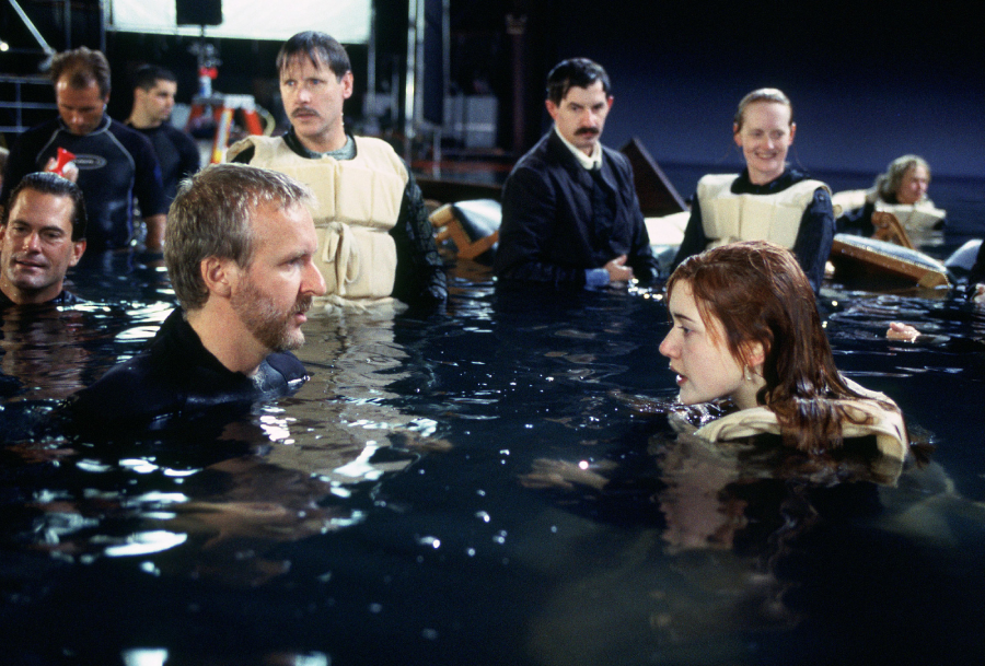 Several people stand in water on a film set, wearing wetsuits and life vests. A bearded man faces a young woman with red hair, while crew members and equipment are visible in the background.