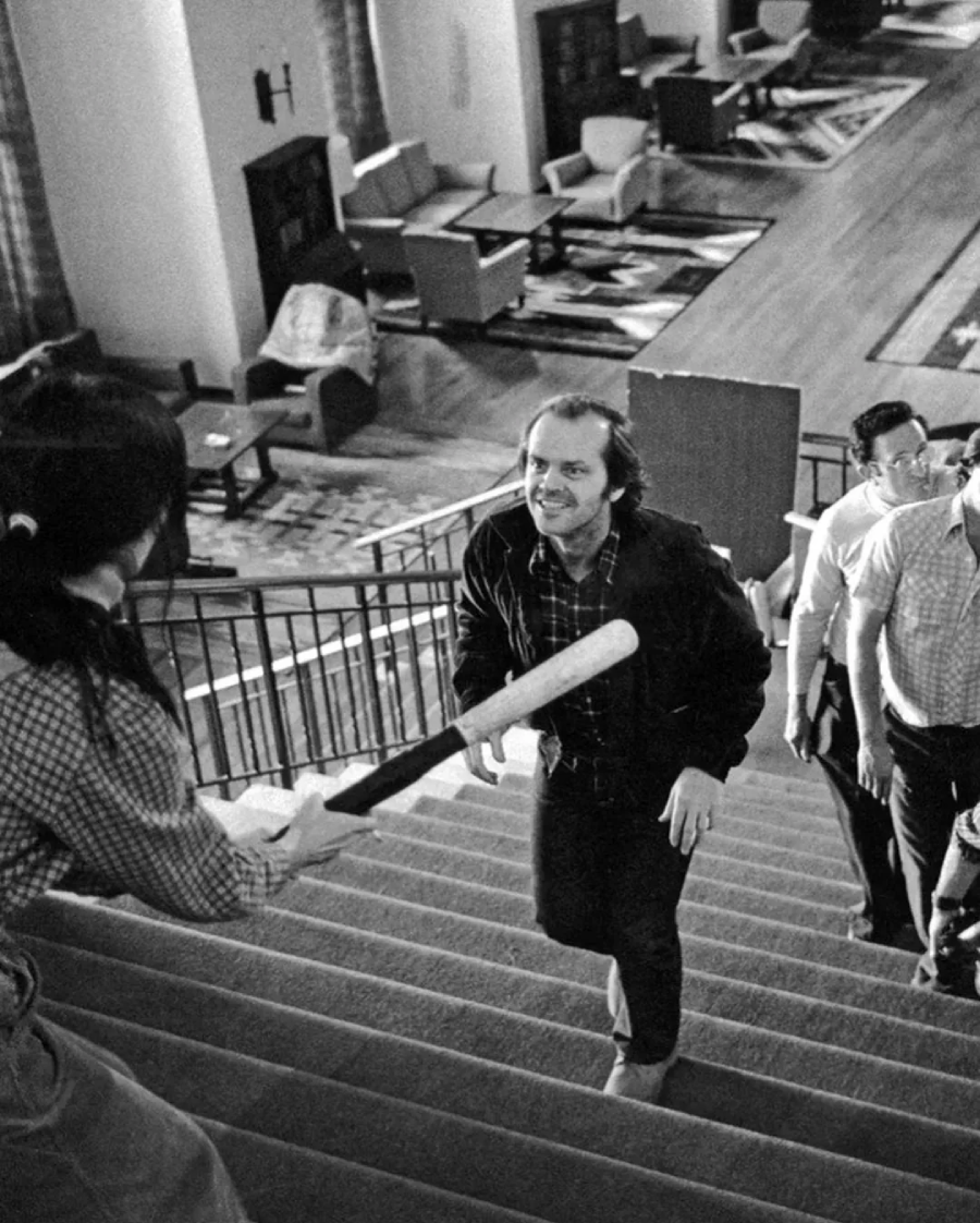 A woman on a staircase holds a baseball bat defensively as a man approaches her, smiling menacingly, while two other men watch from behind in a large, furnished room. The scene is tense and dramatic.