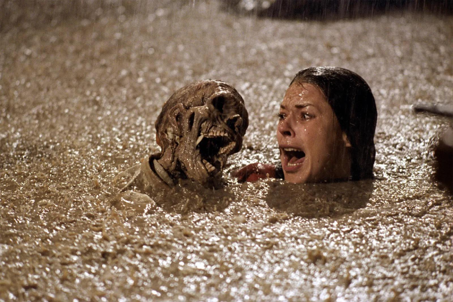A terrified woman screams as she is grabbed by a muddy skeleton emerging from murky water in heavy rain.