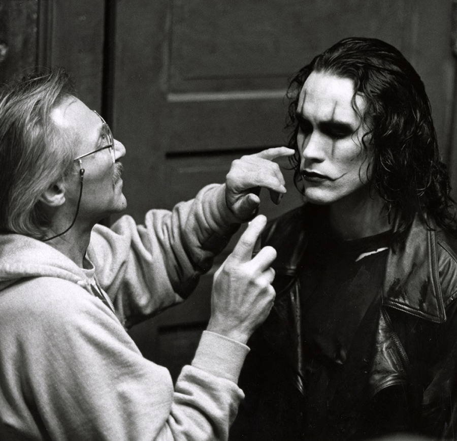 A makeup artist applies makeup to an actor with long dark hair and dramatic face paint, wearing a black leather jacket, standing in front of a wooden door.