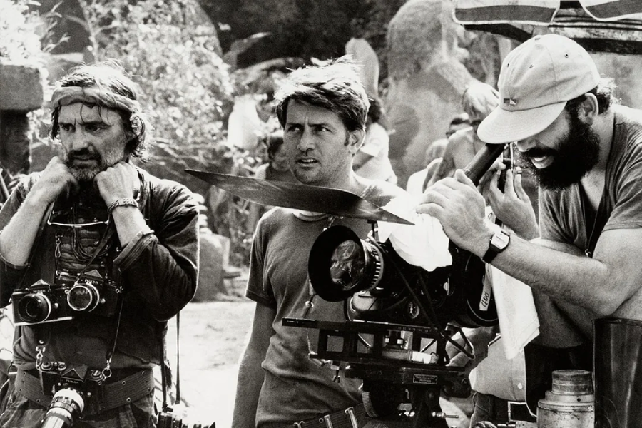 Three men stand on a busy outdoor film set; one wears cameras around his neck, one looks ahead intently, and the third operates a large movie camera, adjusting settings while wearing a cap.