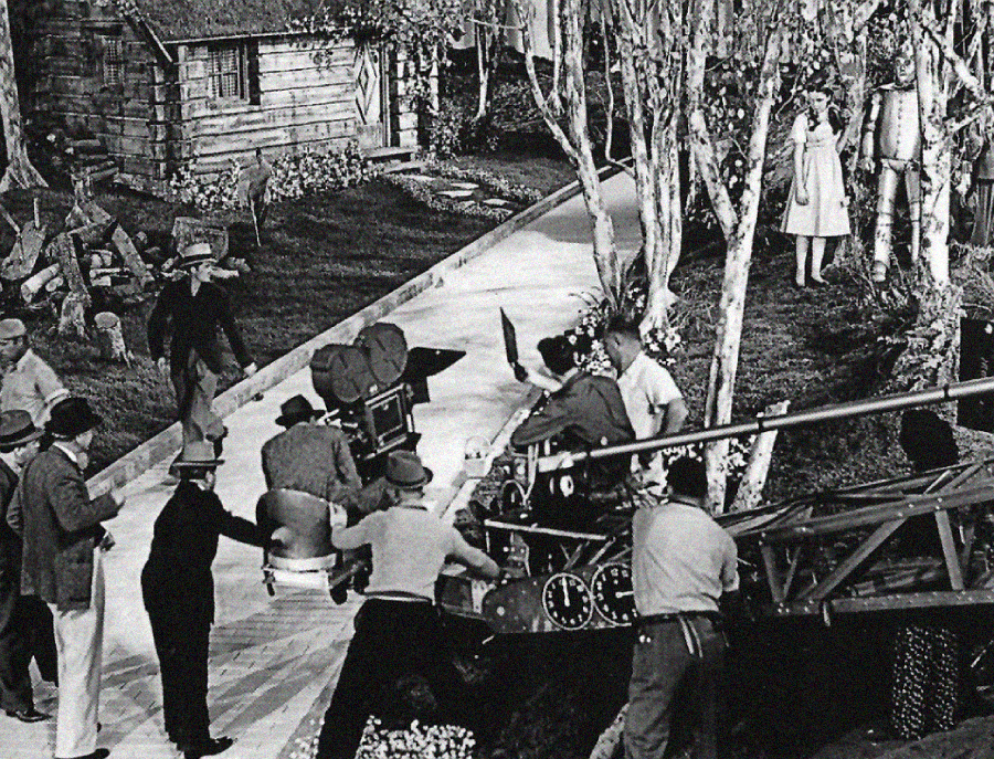 Black-and-white photo of a film crew shooting a scene outdoors. Several people operate a camera on tracks, while actors, including a tin man character and a woman in a dress, stand on a path near trees and a cottage.