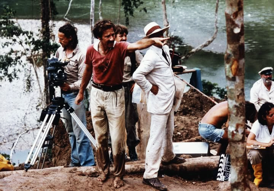 A film crew works by a river in a jungle setting. A man in muddy clothes gestures and points, while another man in a white suit and hat stands beside him. Cameras and crew members are visible around them.