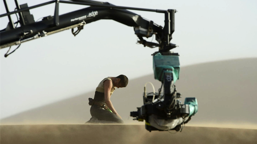 A person kneels on sandy ground in a desert setting, while a large film camera crane captures the scene. The atmosphere is dusty, and sand dunes are visible in the background.