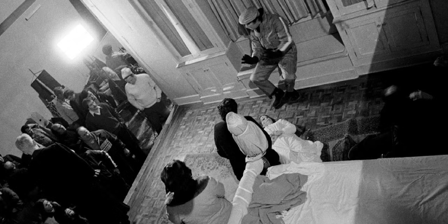 Black and white photo of people gathered in a room, with some standing and others sitting or lying on the floor and bed. A man in uniform sits on a windowsill, observing the scene, while light streams through a window.