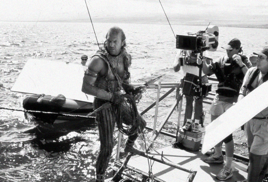 A man in costume stands on a floating platform holding ropes, surrounded by film crew, equipment, and cameras on a body of water during a movie shoot.