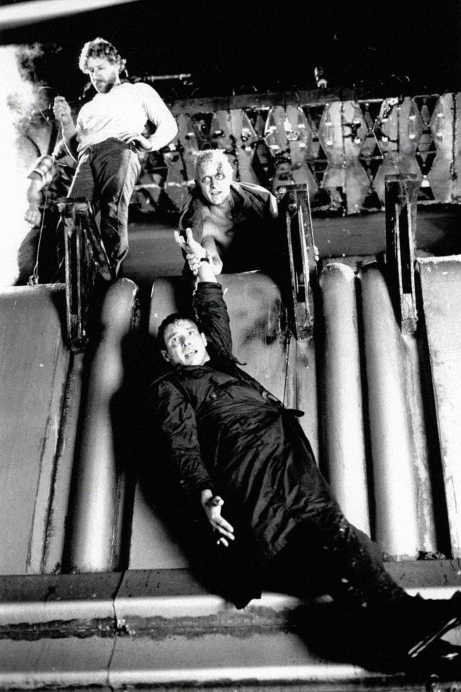 Three men are on the side of a ship; one man hangs from a rail, reaching for help, while another leans over to grab his hand, and a third stands above them holding a rope. The scene is tense and dramatic.