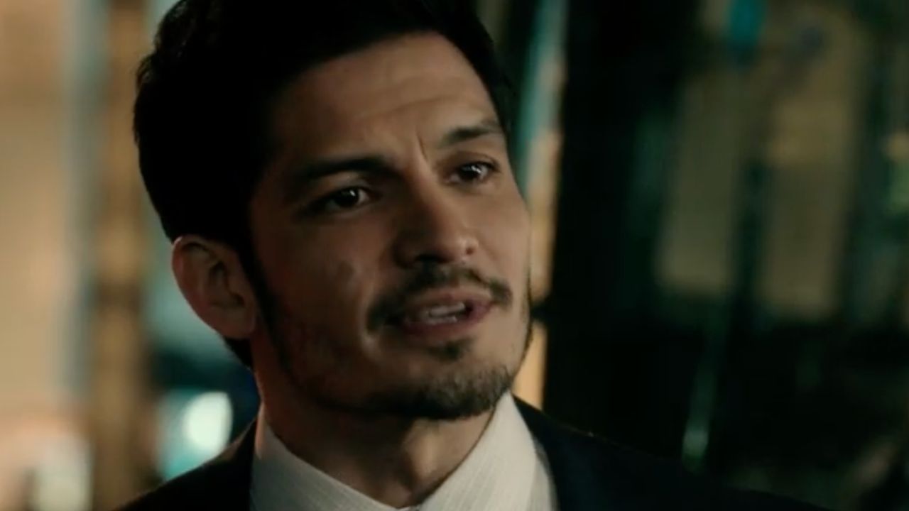 A man with short dark hair, light facial hair, and a serious expression is wearing a suit and white shirt, standing indoors with a blurred background.