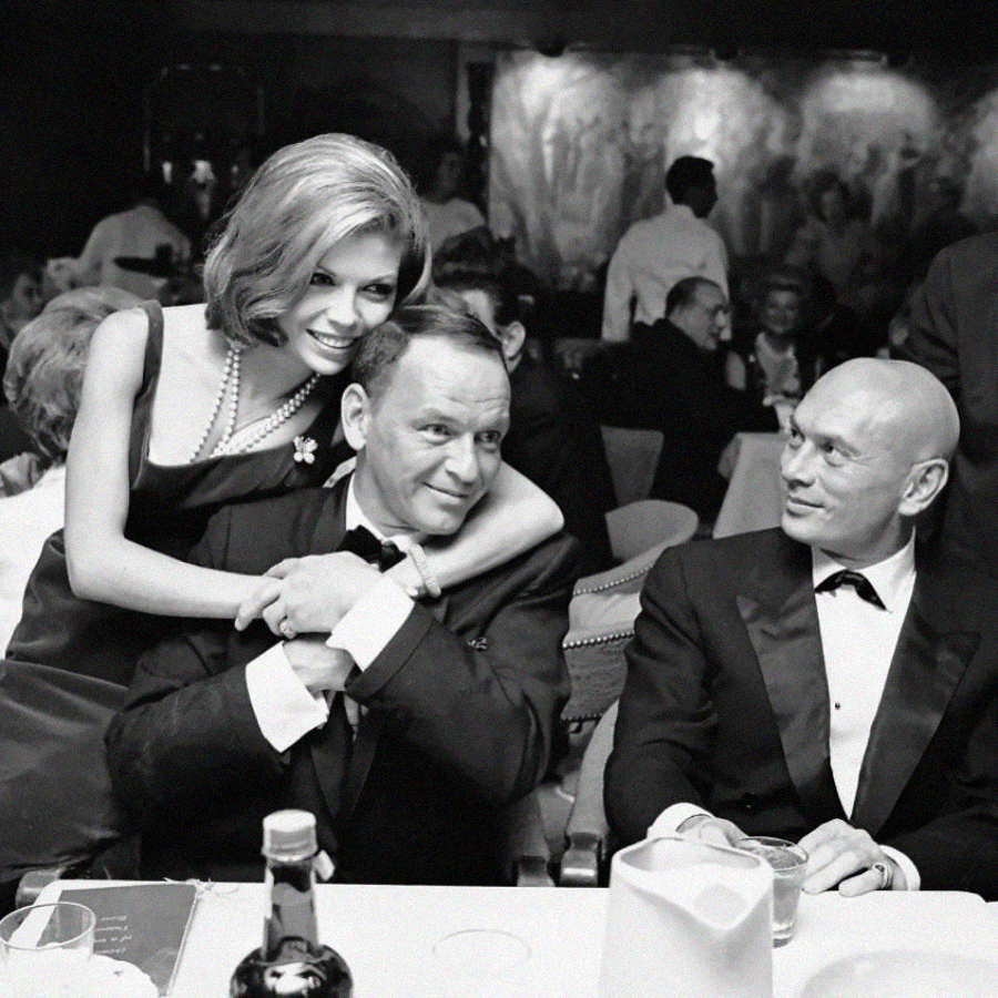 A woman in an evening dress hugs a man in a suit from behind as he sits at a dinner table, while another man in a tuxedo looks on. They appear to be at a lively, upscale restaurant or club.