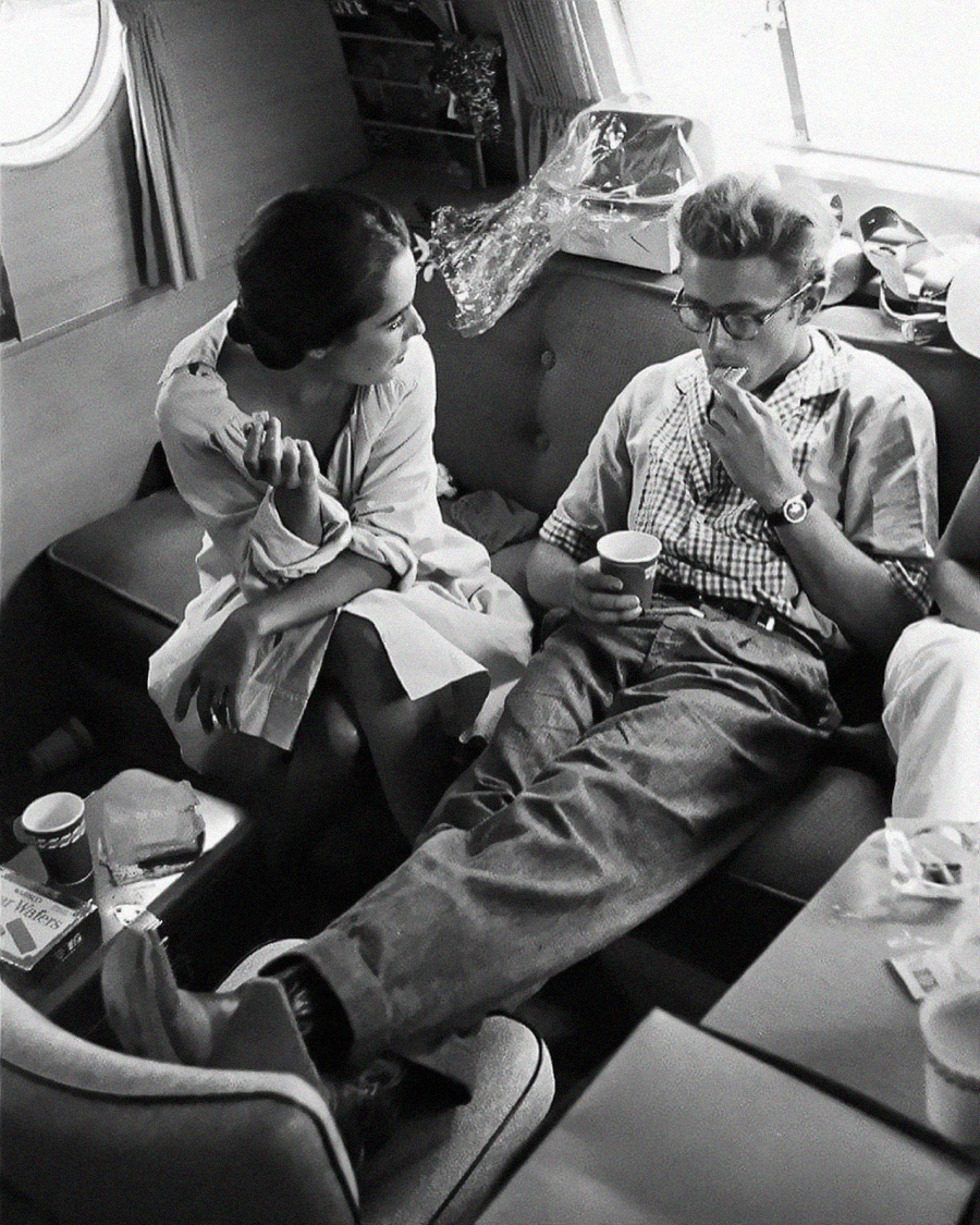 A woman and a man sit closely on a couch inside a cozy cabin. The woman faces the man, speaking and gesturing with her hand, while the man relaxes, legs stretched out, holding a cup and looking thoughtful.