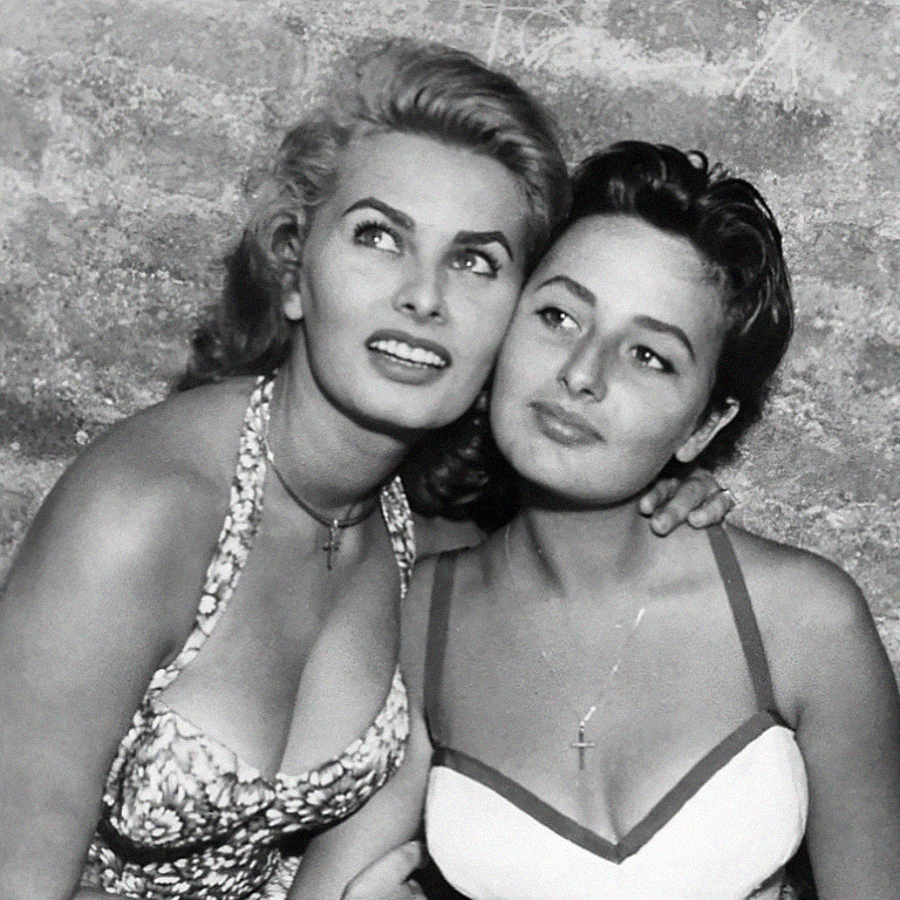 Two women pose closely together, both wearing sleeveless dresses with cross necklaces. They are looking slightly to the side and smiling softly, with a textured wall in the background. The photo is in black and white.