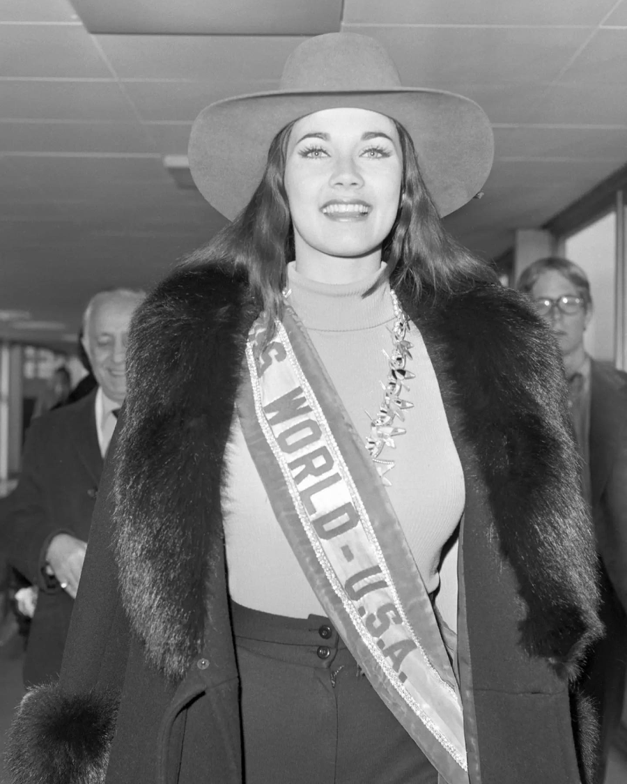 A woman wearing a wide-brimmed hat, fur coat, and a sash reading "MISS WORLD-USA" smiles as she walks indoors, with several people visible in the background. The image is in black and white.