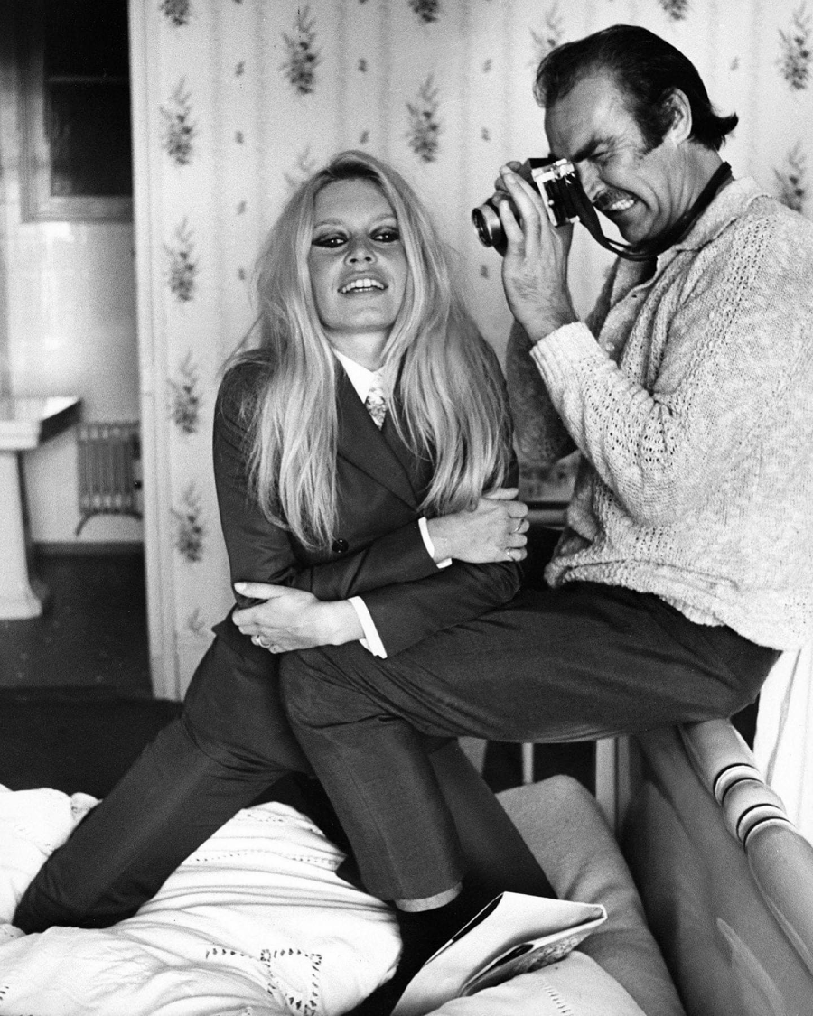 A woman in a suit sits on a bed and smiles playfully as a man in a sweater stands close, photographing her with a camera. The setting appears to be a cozy, patterned room.