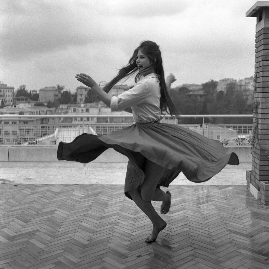 A barefoot woman with long hair twirls joyfully on a tiled terrace, her skirt swirling, with buildings and trees visible in the background under a cloudy sky.