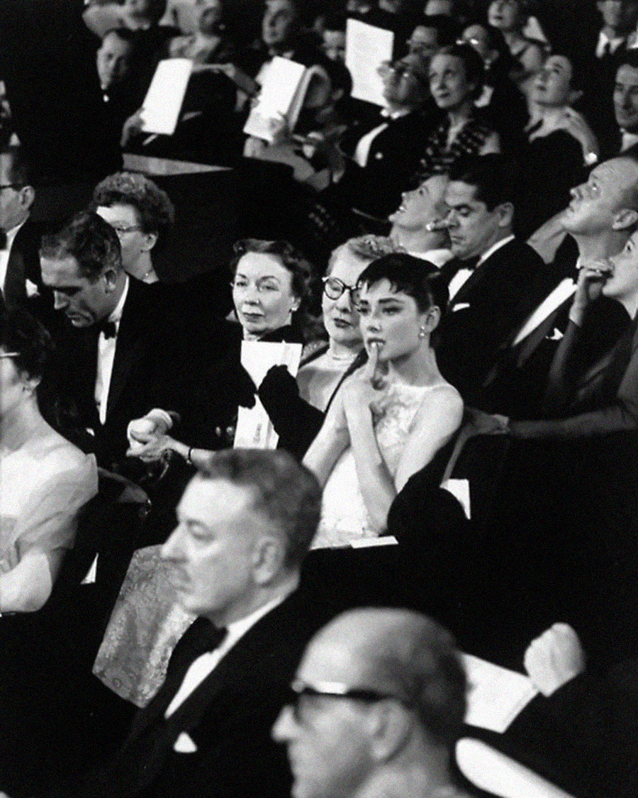A woman in a light dress sits among a crowd of formally dressed people at an event, holding her finger to her lips in a gesture of anticipation or concentration. The audience is mostly focused on the stage.