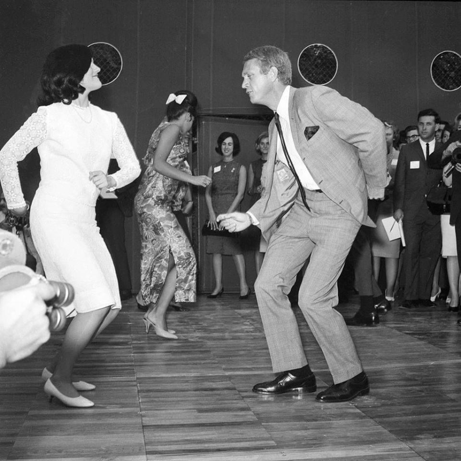 A black-and-white photo of a man in a suit and a woman in a white dress dancing energetically on a wooden floor, surrounded by people watching and smiling in the background.