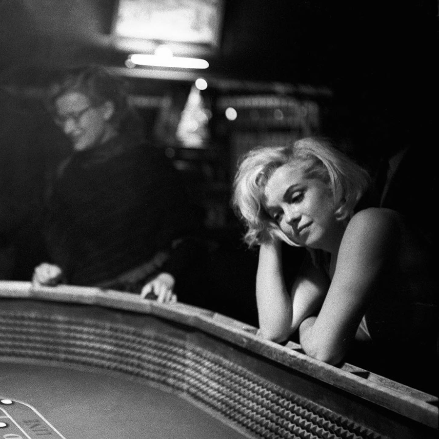 A woman with short blonde hair and a sleeveless dress leans thoughtfully on a casino table, with another person standing nearby in a dimly lit setting.