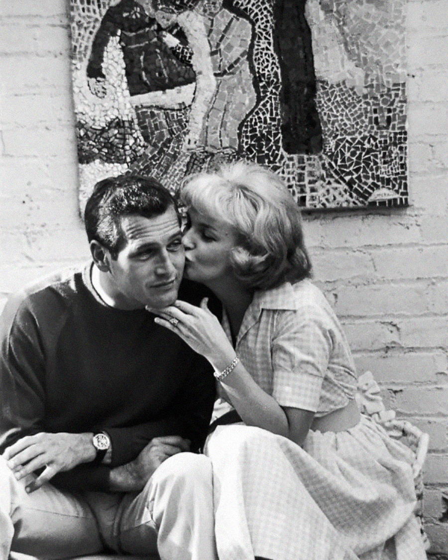 A woman kisses a smiling man on the cheek as they sit on a couch together; a colorful mosaic artwork hangs on the brick wall behind them.