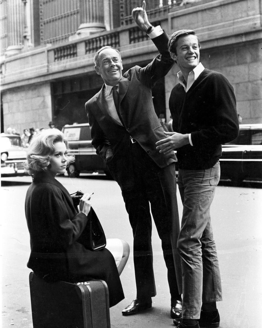 A woman sits on a suitcase smoking, while two men stand nearby on a city street. One man is waving and smiling, and the other, in casual clothes, is laughing. Cars and a large building are visible in the background.