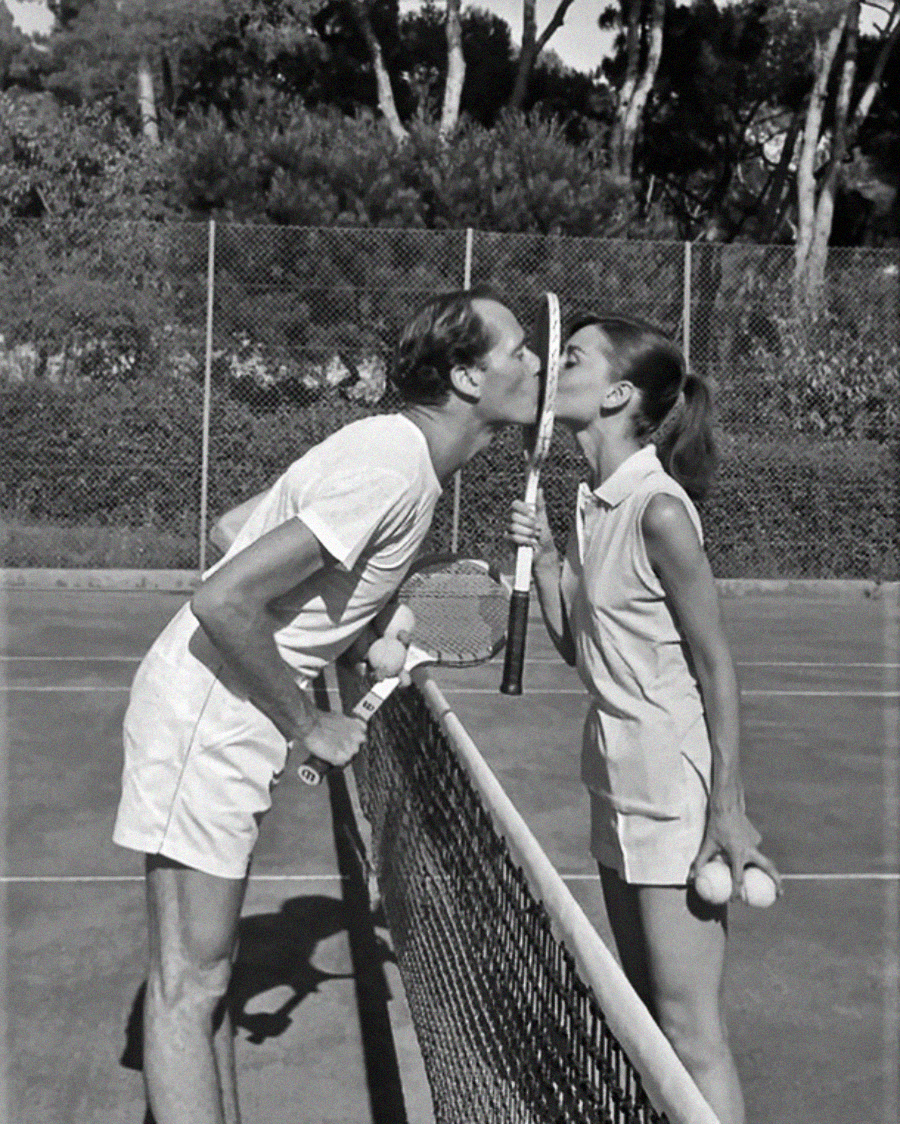 A man and a woman on a tennis court kiss through the net, with a tennis racket held between their faces. Both are dressed in vintage tennis outfits and holding tennis balls. Trees and a chain-link fence are in the background.