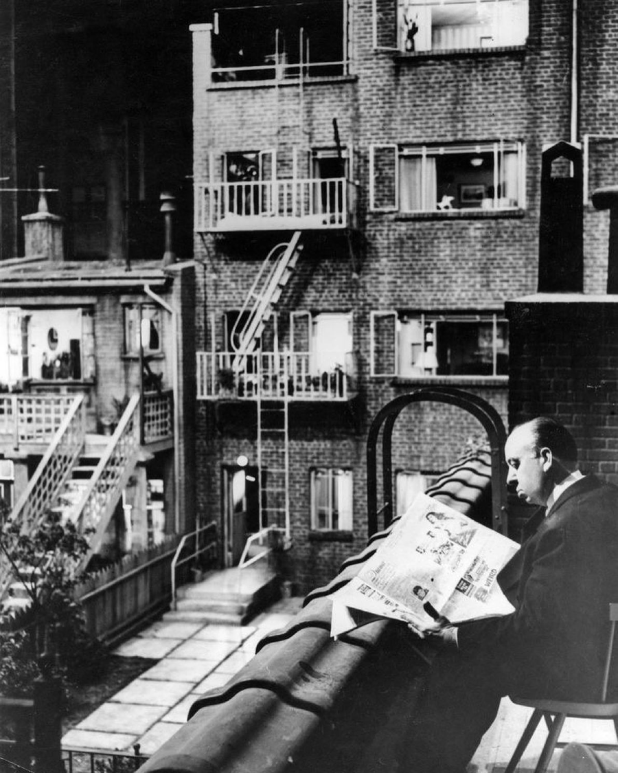 A man sits on a rooftop ledge, reading a newspaper and facing a courtyard with several apartment buildings, balconies, and fire escapes. It appears to be a scene set in an urban residential area.