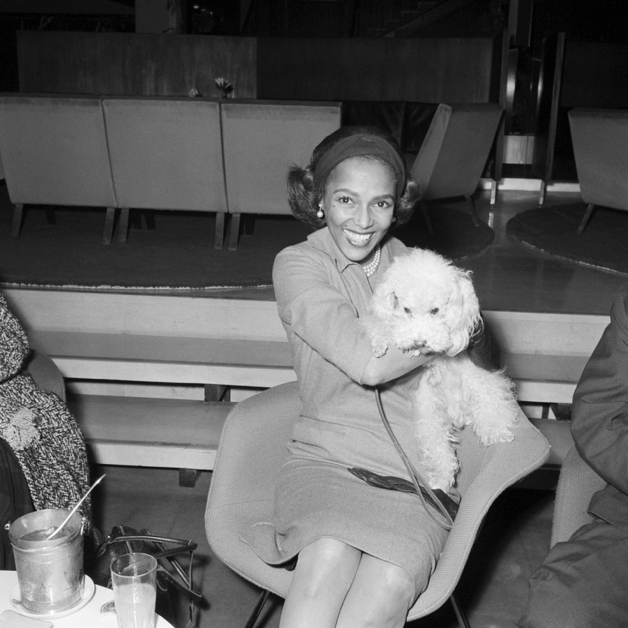 A smiling woman sitting in a chair indoors, holding a fluffy white dog on her lap. There are drinks on the table beside her and modern furniture in the background.