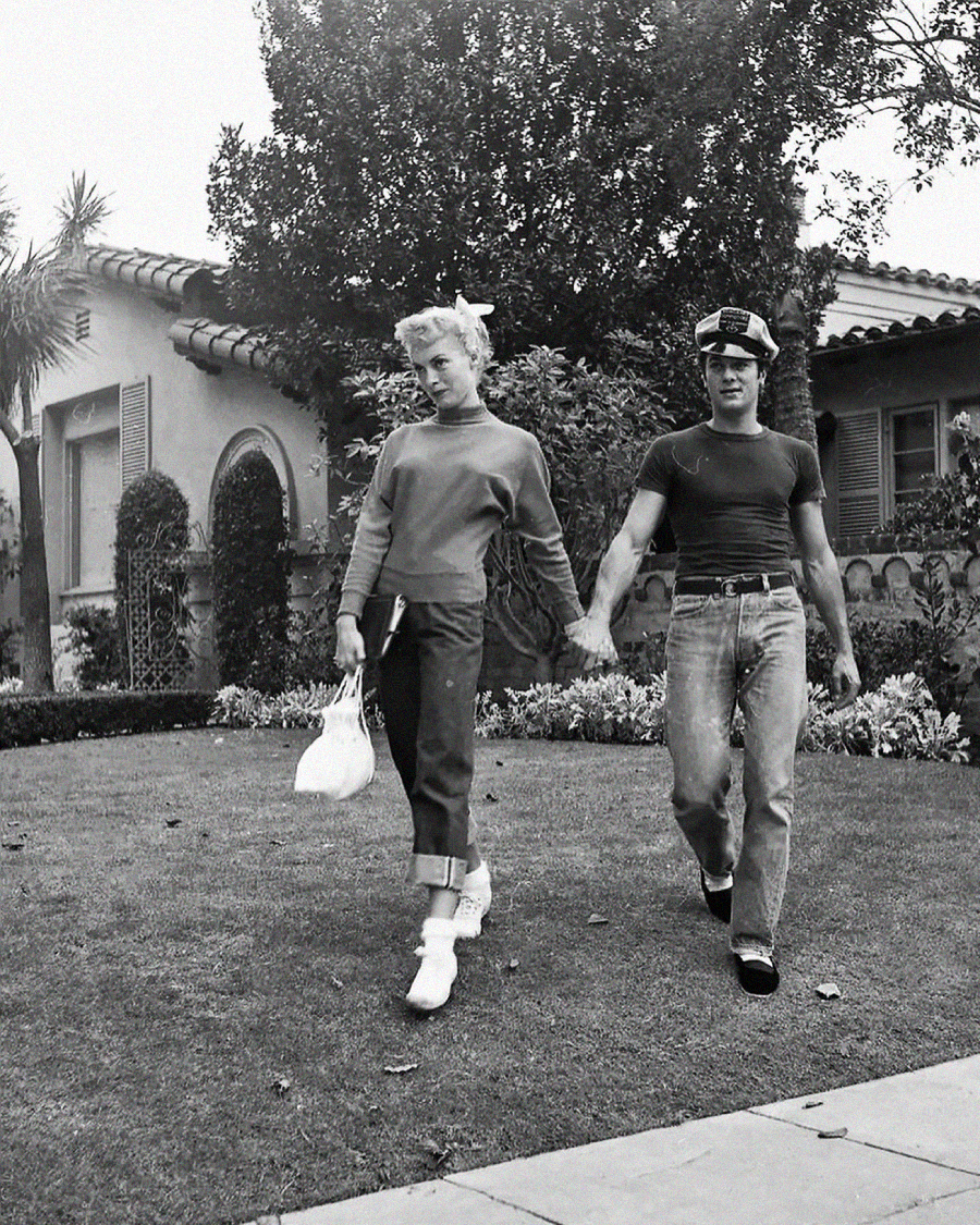 A young woman holding a purse and a young man wearing a cap walk hand in hand on a grassy lawn in front of a house with trees and shrubs, both dressed casually in 1950s-style clothing.