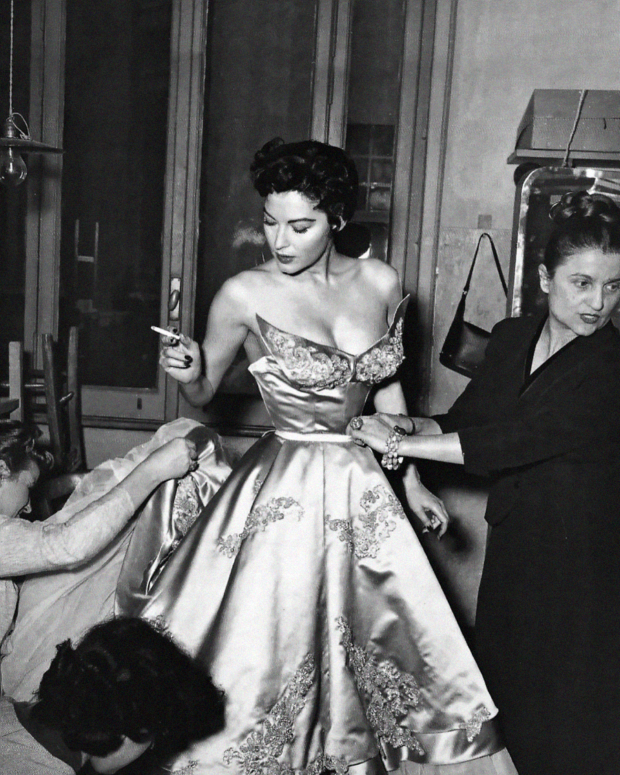 A glamorous woman in an ornate, off-the-shoulder gown is being fitted by three women. She holds a cigarette and stands confidently as assistants adjust her dress in a busy fitting room.