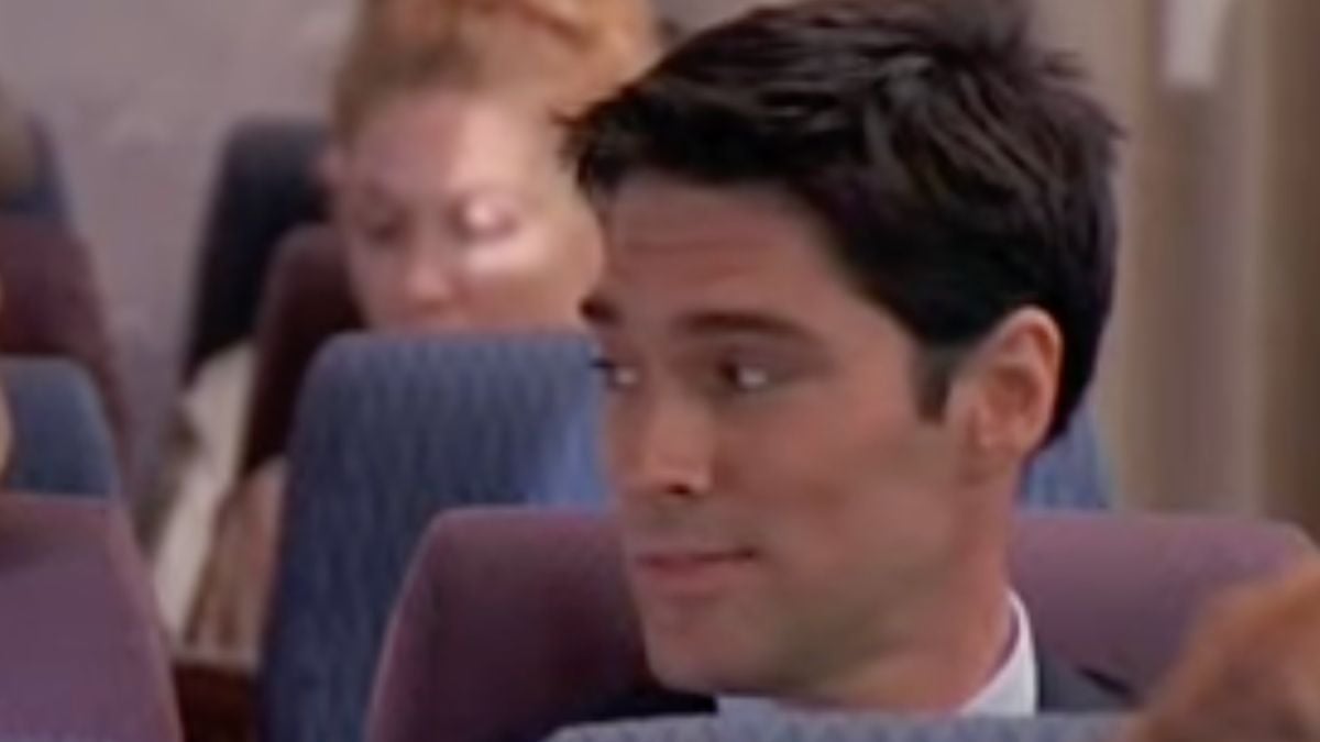 A man with dark hair in a suit sits on an airplane, looking to his left. A woman with reddish hair is seated behind him, slightly out of focus. Both are in typical passenger seats.