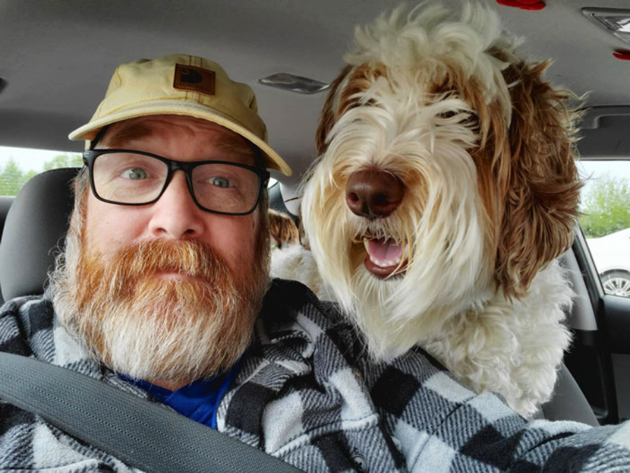 A bearded man wearing glasses and a cap sits in a car, taking a selfie with a large, fluffy dog beside him. The man looks surprised while the dog appears happy, with its mouth slightly open.