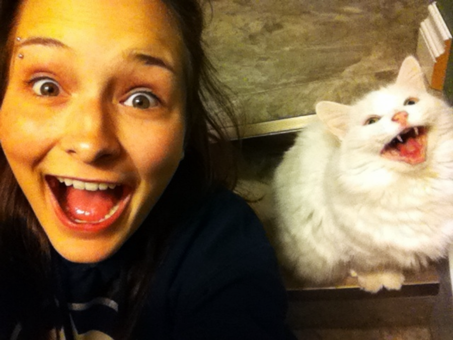 A young woman takes a selfie with a wide-eyed, open-mouthed expression, mirroring a fluffy white cat behind her that appears to be yawning or hissing with its mouth wide open.
