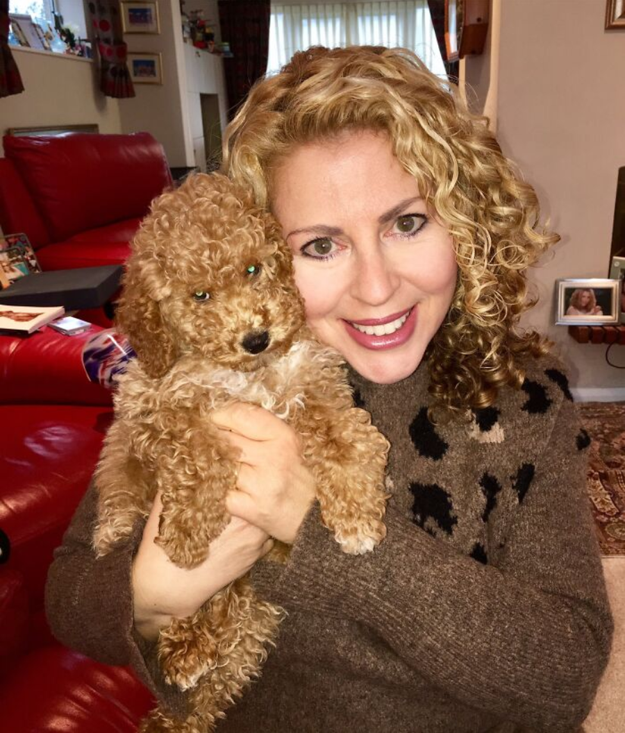 A smiling woman with curly blonde hair holds a small, curly-haired brown dog in a cozy living room with red sofas and framed pictures in the background.