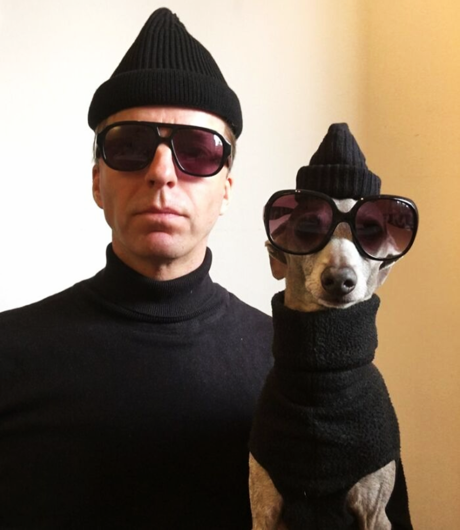 A man and a dog both wear black beanies, sunglasses, and black turtlenecks, posing side by side against a plain background, dressed in matching outfits.