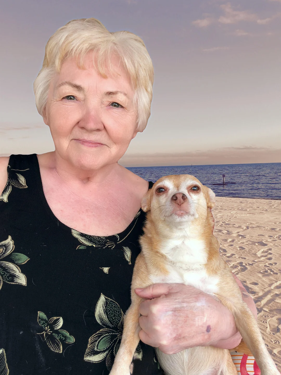 An older woman with short gray hair smiles while holding a small tan dog, standing on a sandy beach with the ocean and sky in the background.