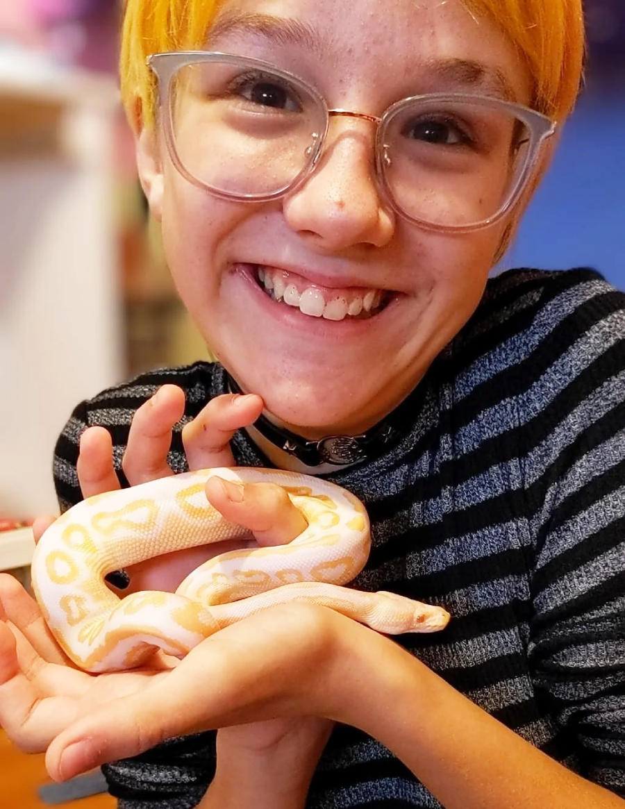 A smiling person with short blond hair and glasses holds a pale yellow and white snake in their hands, wearing a black and gray striped shirt.