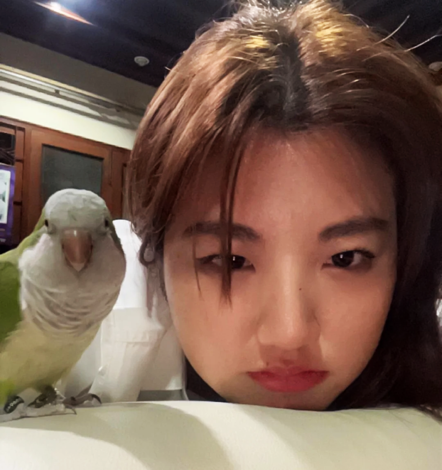 A woman with long brown hair rests her chin on a white surface, looking at the camera with a neutral expression. A green and gray parrot is perched beside her, also facing the camera.