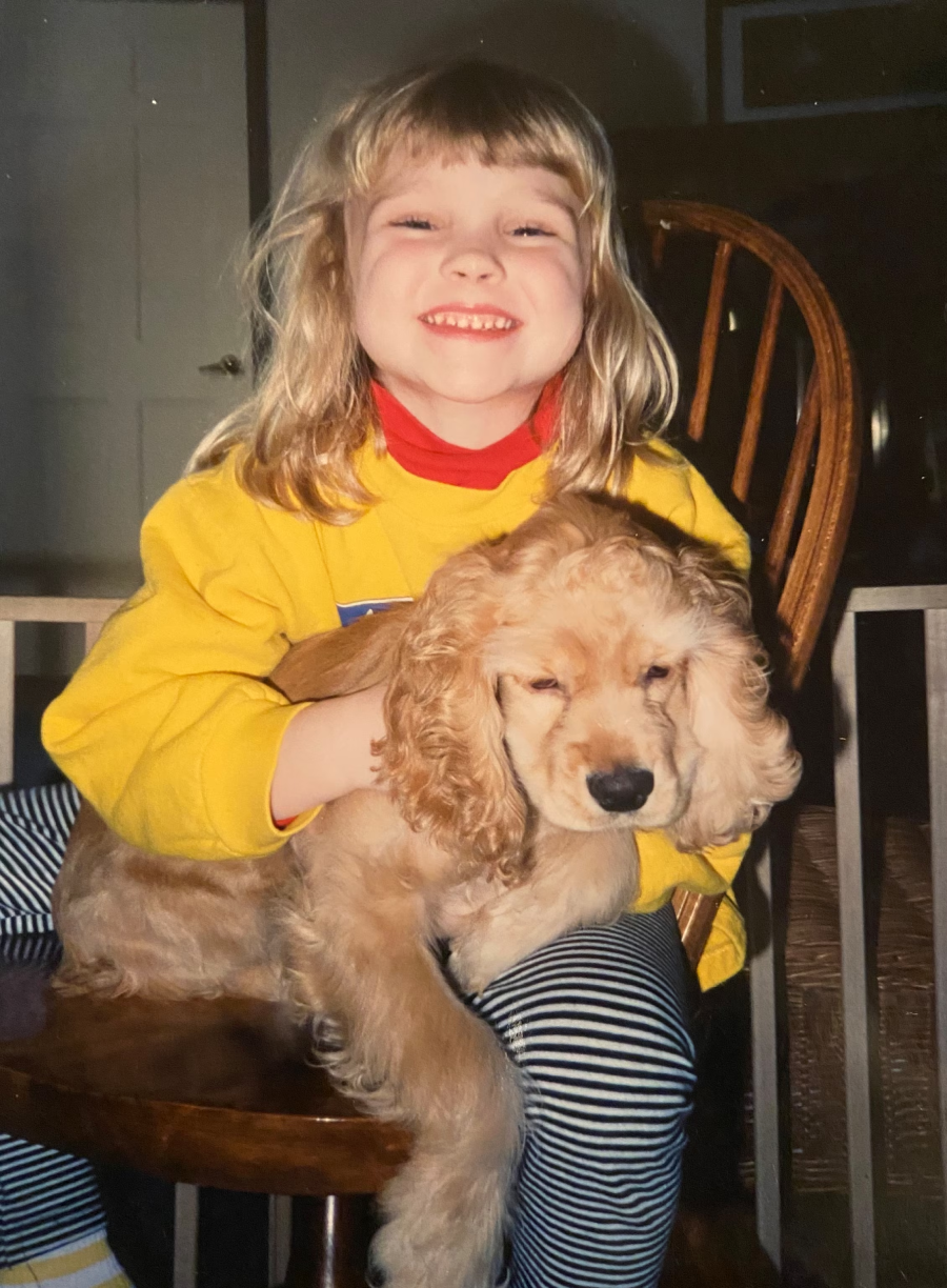 A young child with blonde hair, wearing a yellow sweater and striped pants, sits on a wooden chair and smiles broadly while holding a light brown, floppy-eared puppy in their lap.