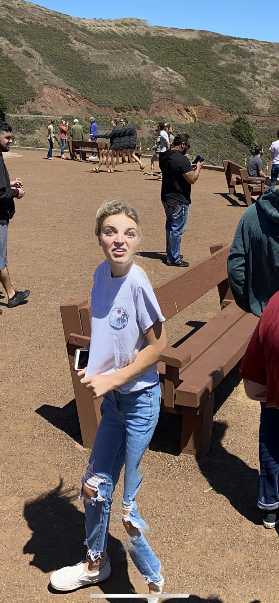 A young woman in a gray t-shirt and ripped jeans stands outdoors on a sunny day, looking up with a surprised expression. She holds a phone and is near brown benches, with people and hills in the background.