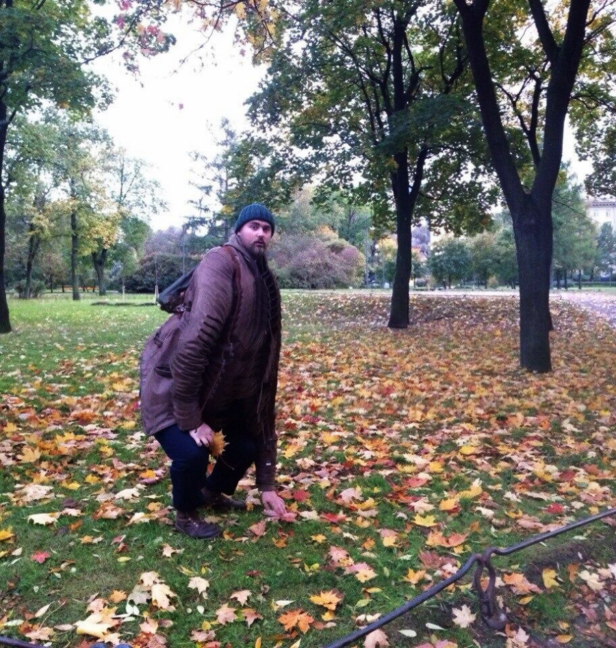 A person in a brown jacket and beanie crouches on grass in a park with autumn leaves scattered on the ground and trees with green and yellow foliage in the background.