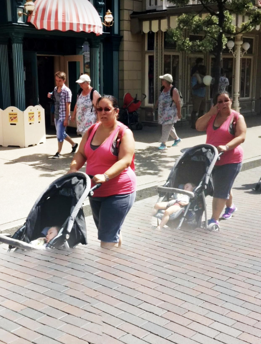 Two women wearing pink tank tops push strollers with young children down a paved street in a busy outdoor shopping area, with other pedestrians and storefronts in the background.