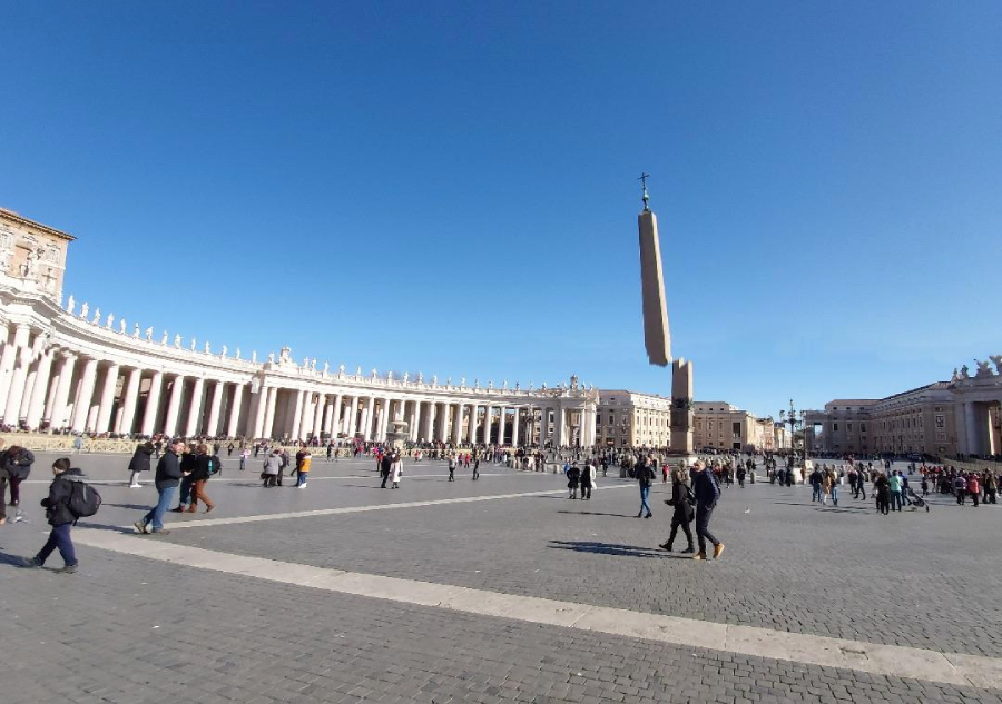 A large plaza with many people walking, bordered by grand colonnades and historic buildings; a tall obelisk stands in the center under a clear blue sky.