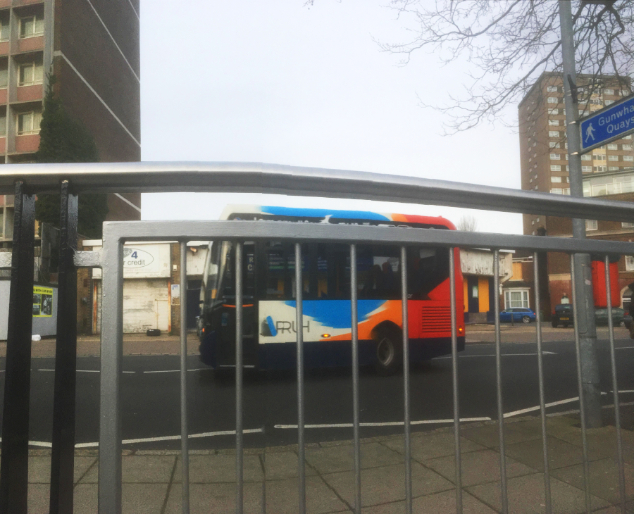 A blue, orange, and white city bus drives past behind a metal railing on a street lined with tall buildings and a road sign reading "Gunwharf Quays." The sky is overcast.