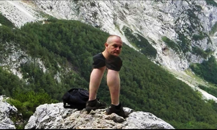 A man standing on a rocky mountain ledge appears distorted, with his legs unusually elongated and his head connected directly to them. A forested mountainside and rocky cliffs are visible in the background.